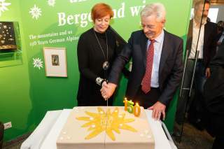 Melanie Grimm und Josef Klenner bei der Jubiläumsfeier 2019. Sie halten gemeinsam ein Messer mit dem sie einen flachen, rechteckigen Kuchen fast durchgeschnitten haben. Der Kuchen ist mit hellem Fondant gedeckt, auf einem goldenen Edelweiß steht 