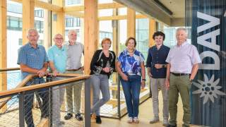 DAV Präsidium in der Bundesgeschäftsstelle. Sieben Personen stehen nebeneinander in einem Treppenhaus aus Holz und Glas. Rechts im Bild an einer Glaswand ist ein Edelweiß und "DAV" vor einem blauen Vorhang auf das Glas geklebt.