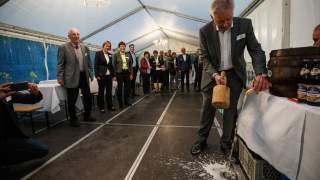 Fassanstich in Bierzelt. Mehrere Personen stehen in einem kleinen Bierzelt. Rechts im Bild schlägt Josef Klenner mit einem Holzhammer den Zapfhahn in ein hölzernes Bierfass. Bierschaum spritzt auf den Boden.