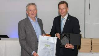 Klenner und Vrijland 2019. Zwei Männer blicken in die Kamera. Der linke hält eine gerahmte Urkunde mit dem Logo der UIAA und des DAV und dem Text "Award of Appreciation", der rechte eine Schatulle mit einem eckigen Glas-Pokal.
