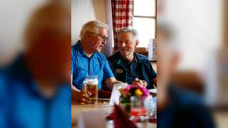 Der Bundespräsident und Josef Klenner bei der Einkehr am Spitzingsee. Zwei Männer sitzen am Tisch und reden. Vor einem steht ein Glas Bier. Vor dem Fenster im Hintergrund hängt ein rot-weiß karierter Vorhang mit Hirschen.