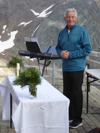 Mann steht auf einer Terrasse, im Hintergrund sind Berge mit Schneeresten zu sehen.