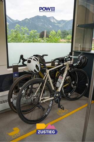 Zwei Gravelbikes stehen am Fahrradstellplatz in einer Regionalbahn. Durch die Zugscheibe sieht man Berge.
