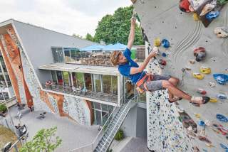 Ein junger Mann klettern an einer überhängenden künstlichen Wand empor. Im Hintergrund ein Dachgarten-Café.