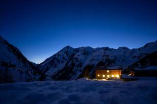 Berghütte am späten Abend, durch die Fenster scheint Licht. Im Hintergrund ist Bergpanorama durch die untergehende Sonne noch leicht zu erkennen.