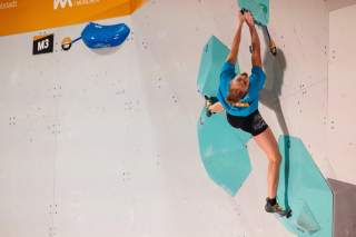 Eine Kletterin hängt an einer künstlichen Kletterwand und greift mit beiden Händen an zwei grüne Klettergriffe, während sie ihre Beine spreizt. Die Wand ist mit einem orangefarbenen Bereich oben und dem Schriftzug 