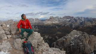 Person in roter Jacke sitzt auf Felsen mit Rucksack neben sich und blickt auf eine weite Berglandschaft.