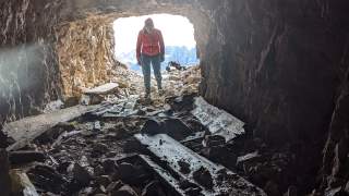 Person mit roter Jacke steht am Eingang einer felsigen Höhle, umgeben von Geröll und alten Holzbrettern, mit Tageslicht von außen.