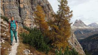 Wanderin auf schmalem Bergpfad, umgeben von Felsen, Nadelbäumen und Berglandschaft im Hintergrund.