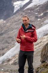 Ein Mann mit Brille und roten Outdoor-Jacke steht vor einer felsigen, teils verschneiten Berglandschaft und gestikuliert mit den Händen.