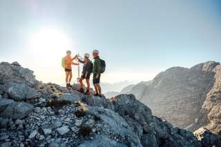 Drei Bergsteiger*innen im Sonnenuntergang auf einem felsigen Gipfel mit Gipfelkreuz, im Hintergrund eine steile Felswand und Gebirgspanorama.