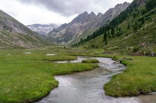 Frei fließender Bach schlängelt sich durch grüne Moorflächen in Tal umgeben von felsigen Gipfeln