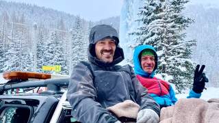 Zwei Männer sitzen zugedeckt im Schneegestöber auf Ladefläche eines Pick-Ups