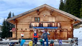 Zwei Menschen sitzen vor Holzhütte, davor liegen Ski im Schnee