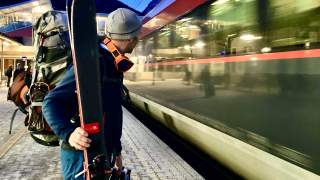 Mann steht mit Ski an Bahngleis, während Zug einfährt