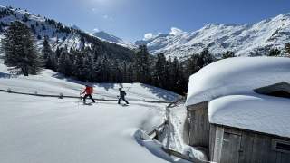 Mann und Frau auf Tourenski passieren eine Holzhütte