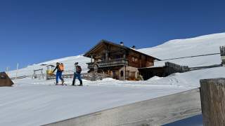 Mann und Frau auf Tourenski passieren ein Holzhaus