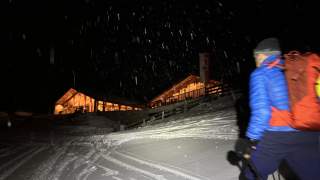 Man mit Rucksack und Tourenski läuft bei Dunkelheit auf eine Berghütte zu.