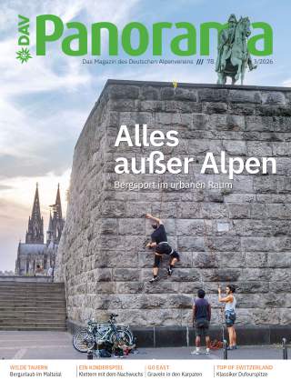 Ein Mann klettert an einer großen Steinmauer in der Stadt, zwei Personen sichern ihn, im Hintergrund ist der Kölner Dom zu sehen.