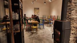 Vier Personen sitzen in einem schlicht eingerichteten Raum mit farbigen Stühlen an einem Tisch, im Vordergrund rechts steht ein Kaminofen vor einer Steinwand.