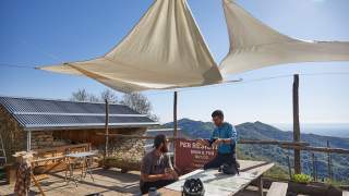 Zwei Männer sitzen an einem Holztisch draußen auf einer Terrasse mit Blick auf bewaldete Hügel, über ihnen spannen sich weiße Sonnensegel. Auf dem Tisch liegen eine Landkarte und zwei Fahrradhelme.