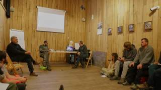 Mehrere Personen sitzen in einem Raum mit Holzwänden in Stuhlkreis-Formation und sehen auf einen Laptop auf einem Tisch, während eine weiße Leinwand an der Wand hängt.