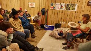 Eine Frau sitzt auf einem Stuhl und zeigt einer Gruppe zuhörender Menschen zwei Filzhüte. Alle befinden sich in einem Raum mit Holzwänden, an denen verschiedene Poster hängen.