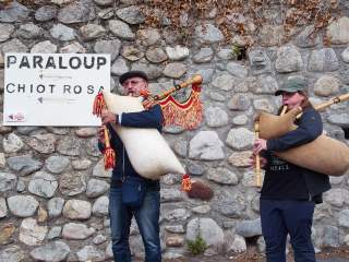 Zwei Personen spielen vor einer Steinmauer Sackpfeife – links ein älterer Mann, rechts eine jüngere Frau, beide mit dem jeweiligen Blasinstrument. An der Wand hängt ein Schild mit den Worten 