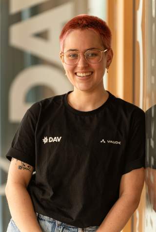 Person mit kurz rasierten pinken Haaren und einer Brille. Die Person trägt ein schwarzes T-Shirt mit dem DAV Logo und eine hellblaue Jeans. Person mit kurz rasierten pinken Haaren und einer Brille. Die Person trägt ein schwarzes T-Shirt mit dem DAV Logo und eine hellblaue Jeans.