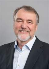 Porträt eines Mannes mit grauem Bart und kurzen Haaren, der in einem hellblauen Hemd und dunklem Sakko lächelnd vor grauem Hintergrund steht.