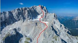 Luftaufnahme eines felsigen Bergmassivs mit einer rot eingezeichneten Aufstiegsroute bis zum Gipfel.