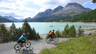 Zwei Männer auf Gravelbikes fahren auf Schotterweg um See, im Hintergrund Berge