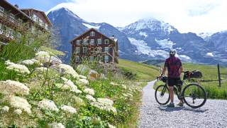 Mann auf Gravelbike steht auf Weg und blickt in Richtung zweier Berggasthäuser, im Hintergrund Berge