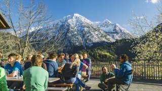 Menschen sitzen auf Terrasse auf Bierbänken, im Hintergrund schneebedeckte Berge