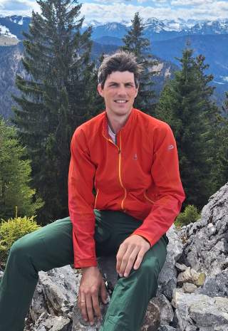 Mann in roter Jacke sitzend auf Felsen. Hinter hm sind Berge zu sehen.