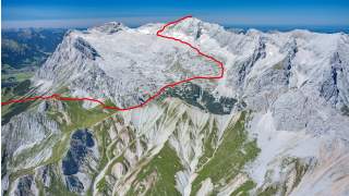 Luftaufnahme eines Gebirges mit grünen und schneebedeckten Flächen, durch die ein roter Linienverlauf als Wanderroute markiert ist.