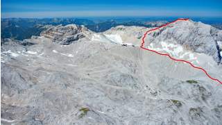 Luftaufnahme eines felsigen Bergmassivs mit vereinzelten Schneefeldern; ein roter Linienverlauf markiert einen Wanderweg über den Grat bis zum Gipfel.