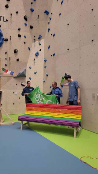 Thomas Michel (Mitte) vom Gay Outdoor Club überreicht die neue Regenbogenbank an Katja Müller.