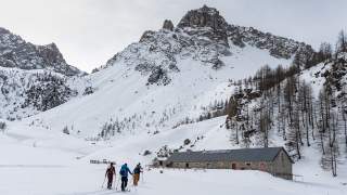Vier Skifahrer in Winterkleidung laufen über eine Schneelandschaft auf eine lange, niedrige Berghütte aus Stein zu, umgeben von schneebedeckten Bergen und kahlen Bäumen.