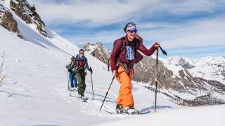 Drei Personen in bunter Winterkleidung beim Skitourengehen auf einer schneebedeckten Berglandschaft unter blauem Himmel.