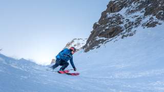Skifahrer in blauer Jacke und Helm fährt schwungvoll einen schneebedeckten Hang mit felsiger Bergkulisse hinunter. Der Himmel ist klar und wolkenlos.