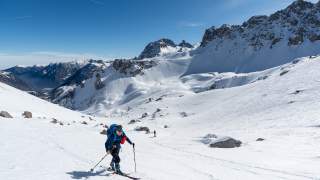 Zwei Skifahrer steigen mit Tourenski und Rucksäcken auf einem schneebedeckten Berg unter klarem blauem Himmel auf, umgeben von schroffen Gipfeln.