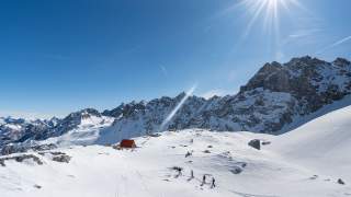 Vier Personen unterwegs auf Skitour unter strahlend blauem Himmel zwischen schneebedeckten Bergen und einem Biwak mit rotem Dach.