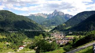 Man sieht die Gemeinde mit ihren vielen kleinen Häsuern und der Kirche, links und rechts bewaldete Hügel und im Hintergrund den Monte Sernio mit seiben felsigen Zacken am Horizont.