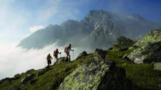 Drei Wandernde steigen den Berg hoch, links sieht man, wie sie die Wolkengrenze hinter sich lassen. Das Wetter ist gut, es ist Sommer, der Himmel ist blau und die Wiesen schön grün.