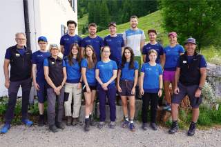 Gruppenfoto von 15 Personen in Outdoor-Bekleidung vor einem Haus mit grüner Berglandschaft im Hintergrund. Die meisten tragen blaue T-Shirts, einige haben Caps oder Sonnenbrillen.