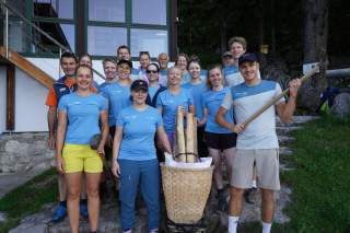 Eine große Gruppe steht vor einem Haus am Waldrand. Sie haben eine Spitzhacke und eine Kraxe dabei und alle tragen hellblaue T-Shirts.