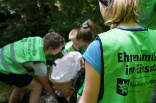 Drei Personen heben eine Kraxe an, im Vordergrund steht eine junge Frau mit dem Rücken zu uns und auf einer grünen Warnweste steht: Ehrenamtlich im Einsatz.