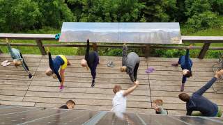 Eine Gruppe junger Menschen sind auf einer Terrasse und machen eine Yogapose. Der Blickwinkel ist dabei von oben.