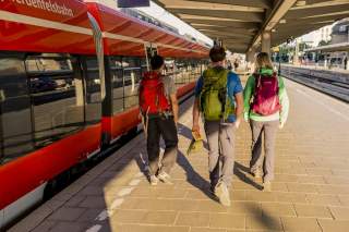 Auf einem sonnenbeschienen Bahnsteig gehen drei Personen an einem roten Regionalzug entlang.
